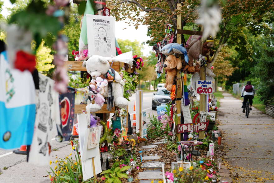 A roadside memorial shows stuffed animals and signs, flags and cards of support.