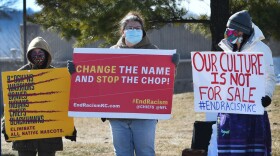 Three people stand outside in an open area. They are holding protest signs. One reads "Change the name and stop the chop!" Another reads "Our culture is not for sale."