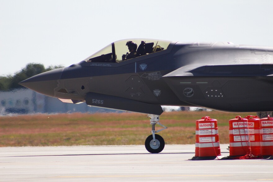 A photo of a person in a cockpit of a fighter plane on a runway with warning signs. 