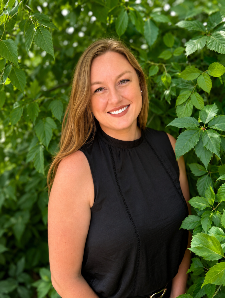A woman wearign a black top standing in front of a lot of green leaves.