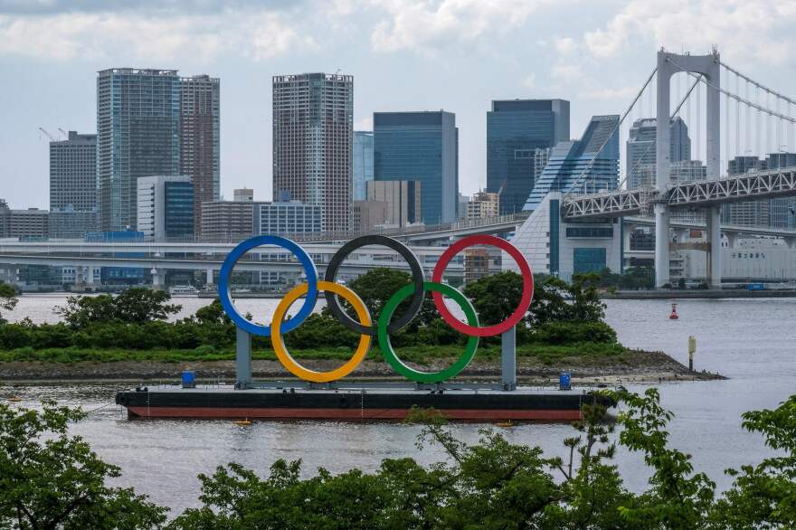 A large statue of the five colorful Olympic rings sits atop the Odaiba waterfront in Tokyo, against the grey backdrop of a bridge and high-rise buildings.