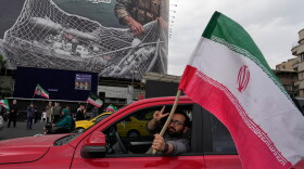 A man flashes a victory sign as he carries an Iranian flag in front of an anti-U.S. billboard depicting the American aircrafts into the Iranian armed forces fishing net with signs that read in Farsi: "The Strait of Hormuz will remain closed, The entire Persian Gulf is our hunting ground," at the Eqelab-e-Eslami, or Islamic Revolution Square in downtown Tehran, Iran, Monday, April 13, 2026.