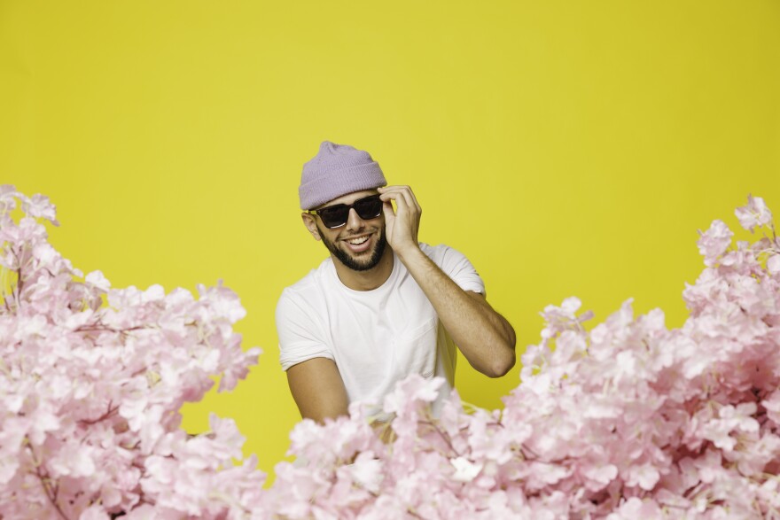 An artistic headshot of Joe Hertler. Light pink flowers crowd the foreground. Hertler sits behind them, wearing a matching pink beanie. One hand rest in his lap, and another holds onto the frame of the sunglasses he's wearing. His plain white t-shirt stands out against the neon yellow background. 