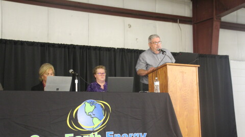 A business executive speaks at a podium next to a table set up for a public meeting. 