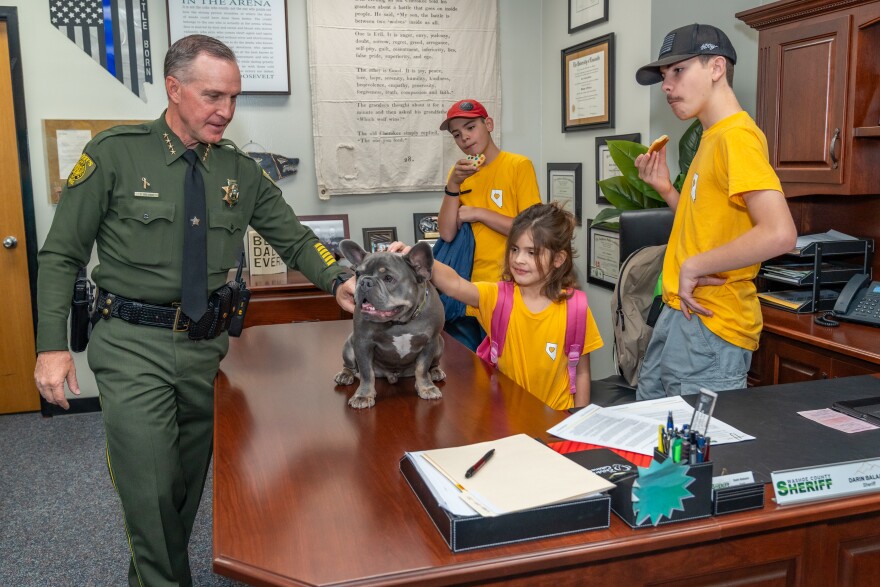 Chance sits on Sheriff Balaam's desk while receiving pets from children from Northern Nevada Children's Cancer Foundation