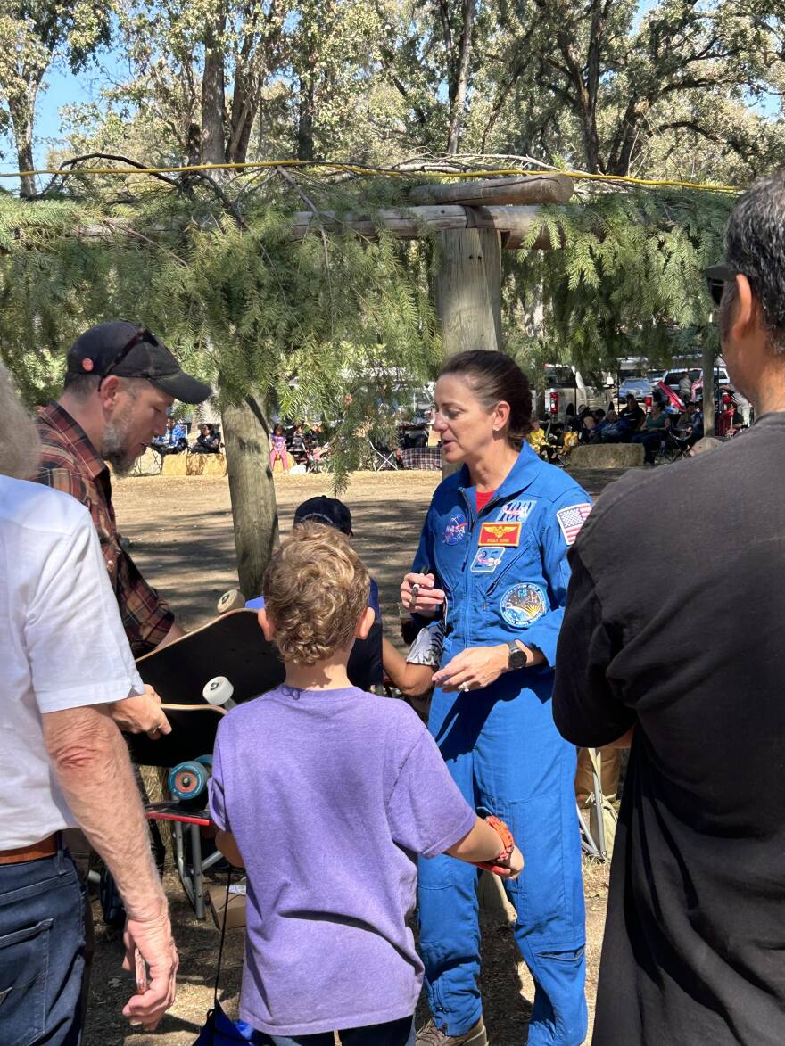 A woman in a blue NASA uniform speaks to people in a park with trees and cars in the background.