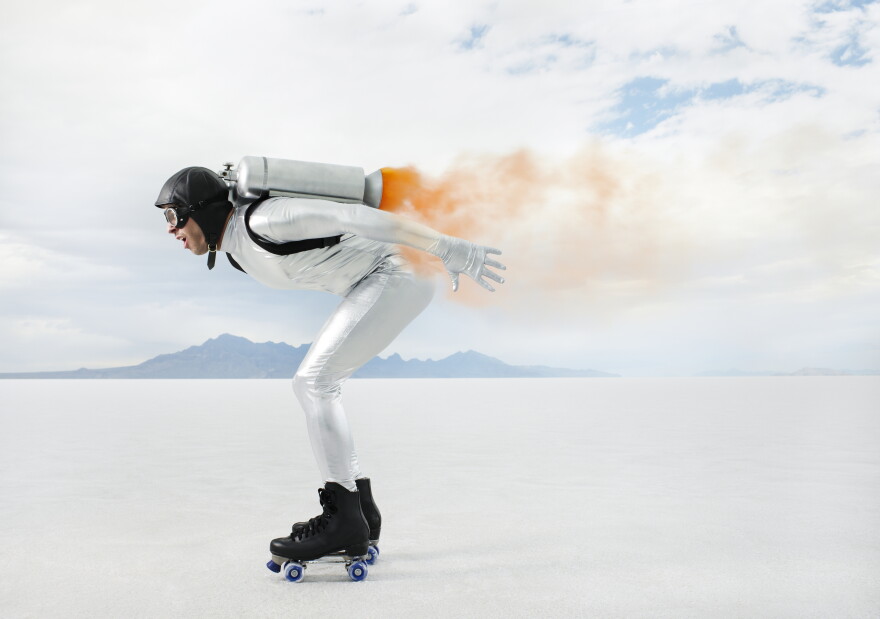 FILE: A wide, horizontal shot of a person wearing a white jumpsuit, black roller skates, and a jetpack, gliding across a vast, flat salt plain with mountains in the background under a cloudy sky. The person is in a crouched position with arms outstretched, surrounded by a cloud of orange smoke emitted from the jetpack. The scene is bathed in soft, diffused daylight, capturing a sense of adventure and innovation.