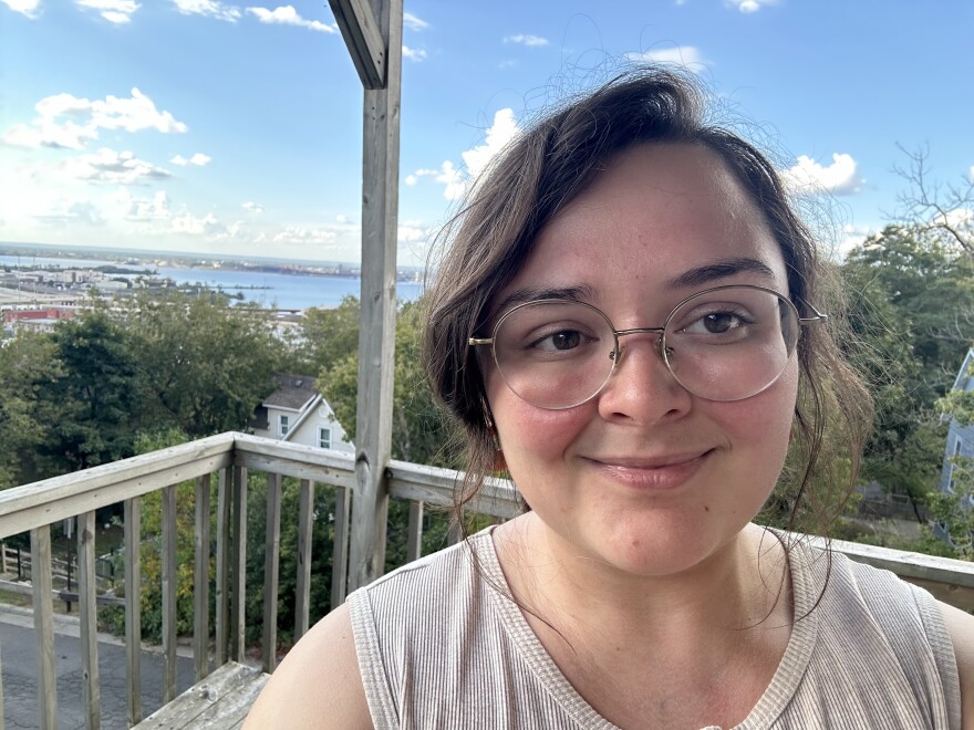 A smiling young woman seen from the shoulders up on a deck with blue sky, trees, and a view of a harbor in the background.
