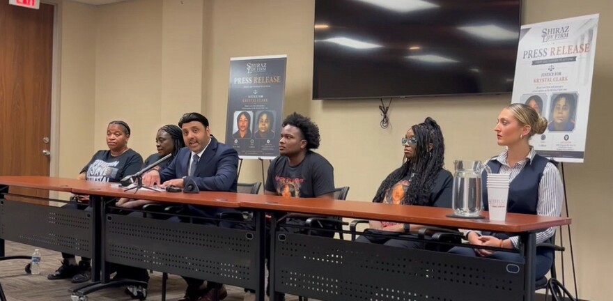Attorney Shiraz Khan sits by Clark's mother to his right and Clark's son to his left. Large promotional posters from the Shiraz Law Firm bearing the headline "Press Release: Justice for Krystal Clark" stand in the background.