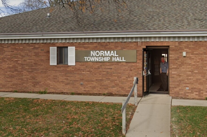 A brick building with the sign "Normal Township Hall" above the entrance. A person is partially visible standing in the open doorway. The front has a sidewalk and some grass with fallen leaves.