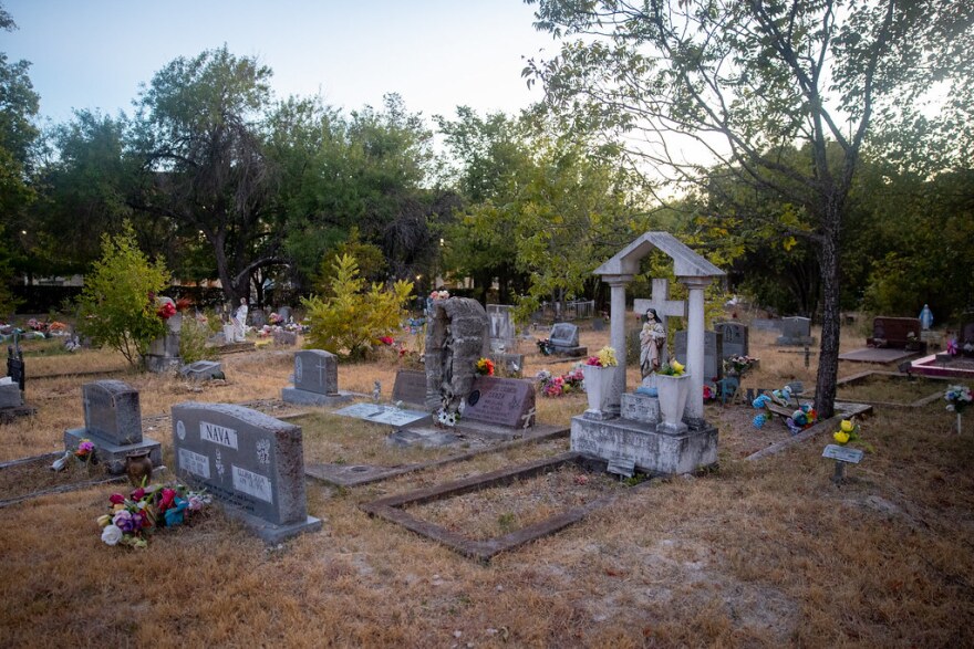 A wide view of several rows of graves at the cemetery. Most prominent is a grave with a large headstone that includes a statue of Jesus Christ in front of a stone cross. Flowers of various colors are placed at most plots. A row of tall trees lines the background. 