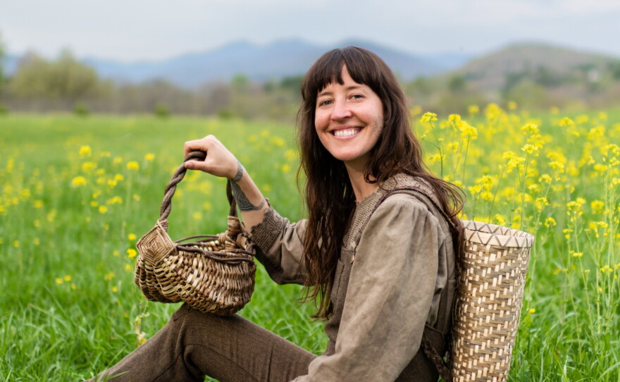 A lady sits in a field smiling with kudzu baskets and a white oak pack basket.