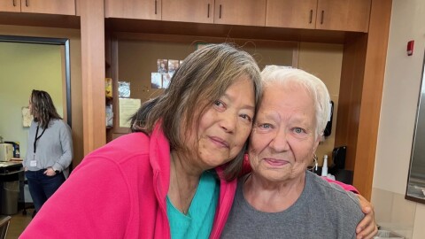 Two women stand close together, one holding a plate with bread and a bowl of soup.