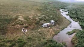 A photograph of a remote, grassy landscape with a few small shacks on it, near a river.