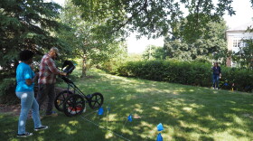 Stacey Camp, is holding up her index finger, pointing towards the ground penetrating radar, next to several small flags which establish the detection grid for the radar device outside on a patch of grass