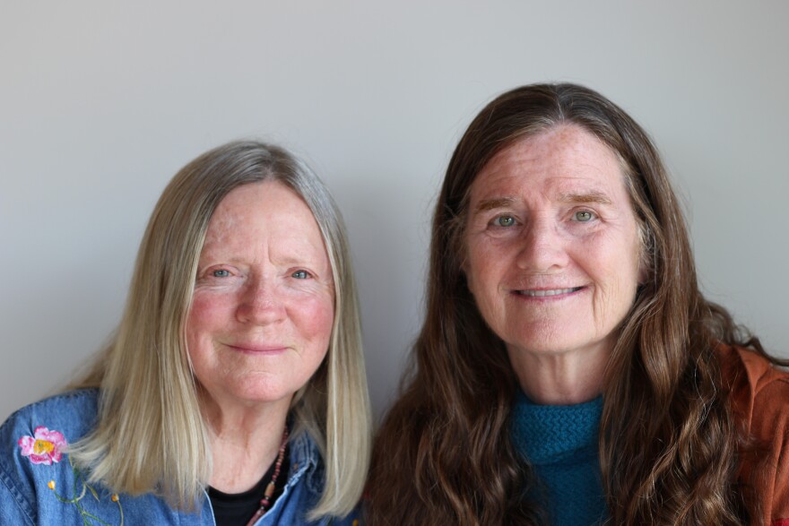 Kathleen Kavarra-Corr and Bernadette Kane-Cole side by side, smile into the camera.  Dr. Kavarra-Corr has straight, shoulder length blonde hair, parted in the middle.  She wears a blue denim  machine embroidered button-up shirt with floral designs, partially unbuttoned over a black tee shirt, and a wooden beaded necklace.   Bernadette Kane-Cole has  long, wavy brown hair.   Her smile shows her teeth.  She wears a blue textured sweater with a rust colored hoodie.