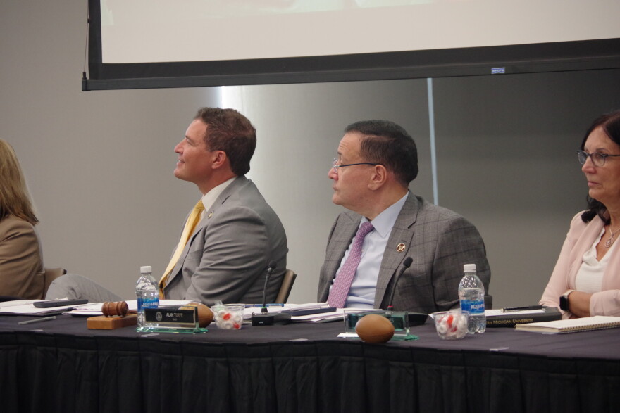 At a black table, Russ Kavalhuna sits at an angle, smiling at someone off camera. He is wearing a grey suit with a yellow tie. Next to him, Alan Turfe also looks off camera. He is wearing a grey suit with a purple tie. Next to them, board secretary Stacey Thinnes is pictured in a light pink suit with arms crossed. 