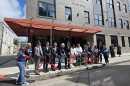 A line of people holding large pairs of scissors cut a red ribbon in front of an apartment building