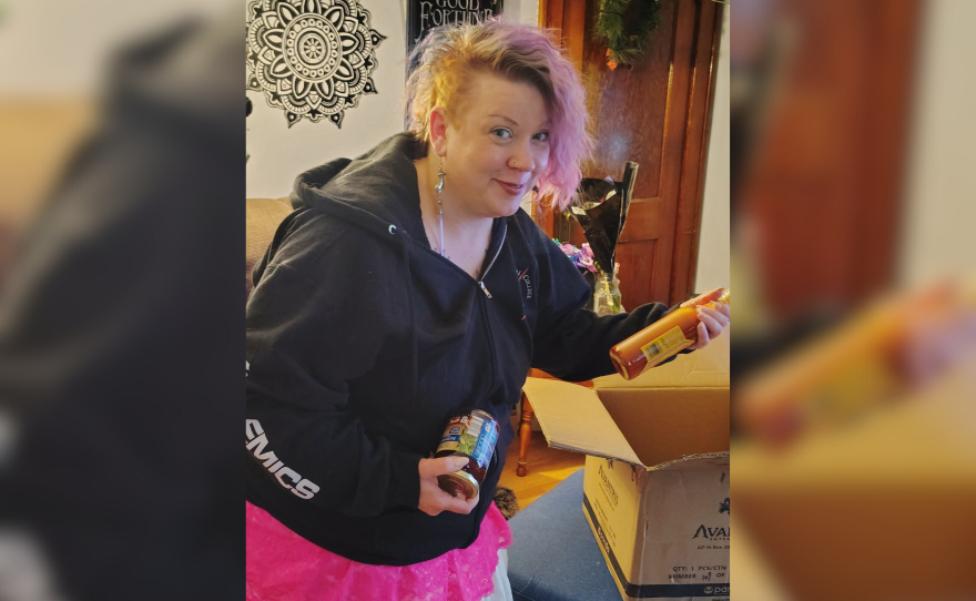 A woman in a black sweatshirt and pink skirt packs food items into a box.