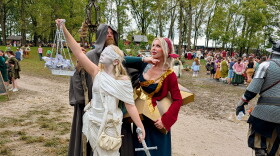Three people dressed in costumes pose together on a muddy road. The first person is wearing all white and is holding a set of scales and a sword. The second person is wearing a long grey hooded robe and carries a staff. The third person is wearing a red and blue outfit with a gold star structure around her waist. 