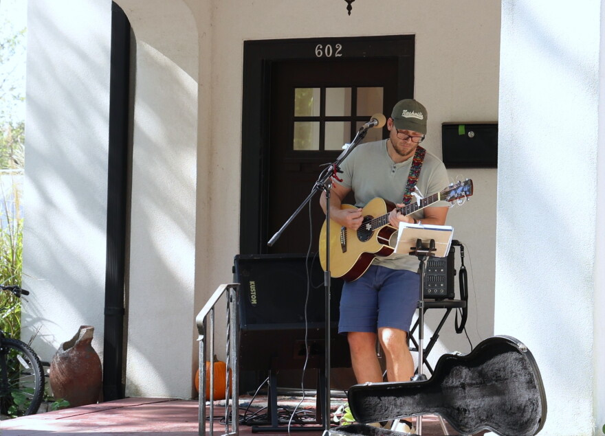 Trevor C. Smith performs at the 10th annual North Heights Porchfest in Joplin on Oct. 10, 2026. He's on a front porch, standing behind a microphone and in front of a speaker. He's playing an acoustic guitar with the case open in front of him.