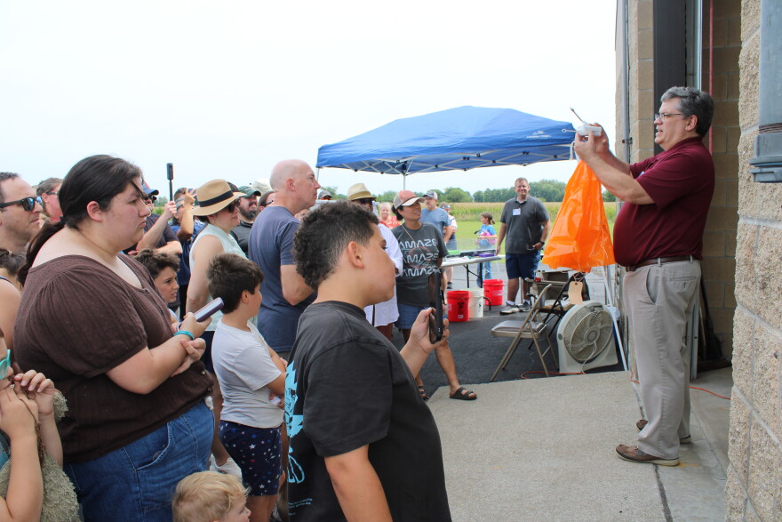 A man stands in front of a crowd holding a weather instrument. He is explaining to the crowd weather balloons work right before he launches one.