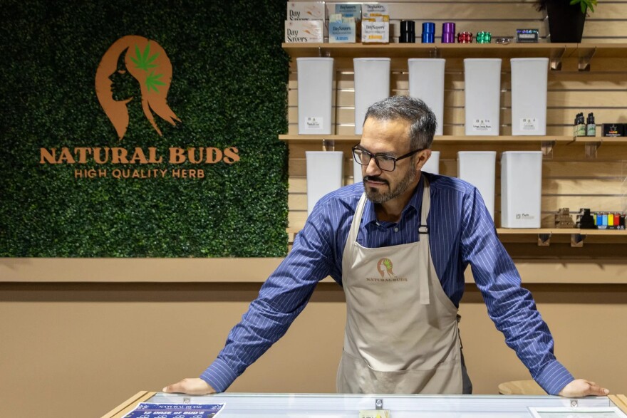 A man in an apron stands at a counter and a sign behind him reads "Natural Buds High Quality Herb."