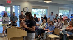 A tattooed white woman stands at a podium and reads a speech. Behind her is a crowd of people -- mostly white, many of them wearing blue. 