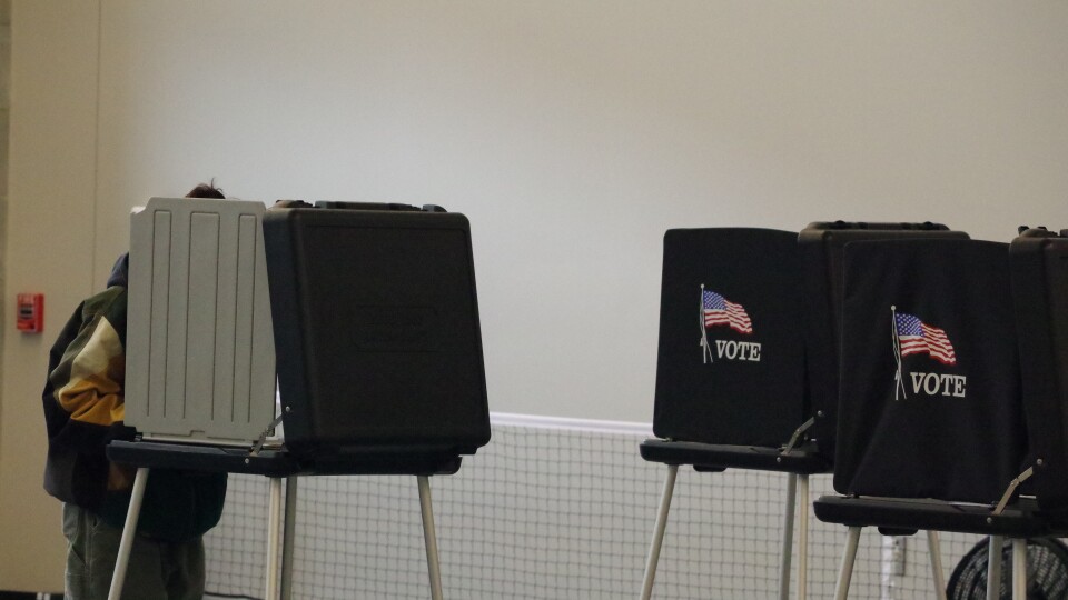 A person in louse-fitting green pants stands at a black voting booth, only parts of their figure visible. Two other black voting booths can be seen to the right, both displaying an emblem with the word Vote underneath an American Flag. 