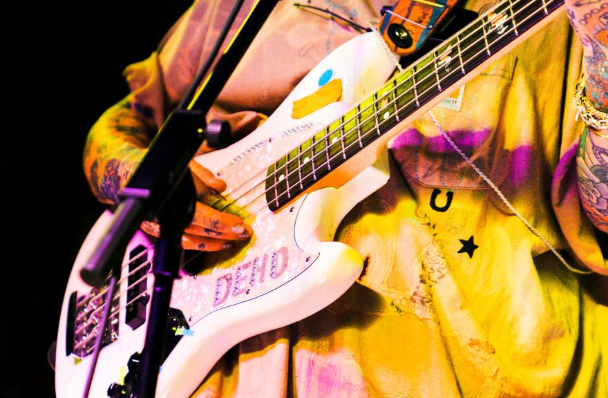 The torso of a person wearing a yellow shirt is shown under a white electric guitar with stickers reading "Dehd."