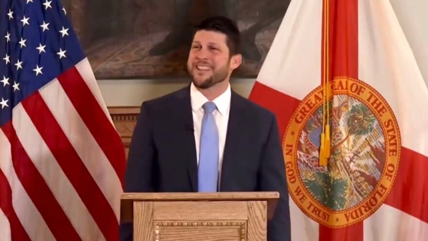A man with dark brown hair and a short beard wearing a dark suit and light blue tie stands behind a podium addressing a crowd. A Florida and US flag stand behind him. 