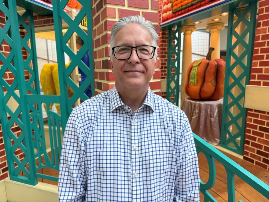 A man wearing glasses with gray hair and a white shirt with thin-lined blue squares is smiling and standing in front of a big art installation depicting the Iowa State Fair.