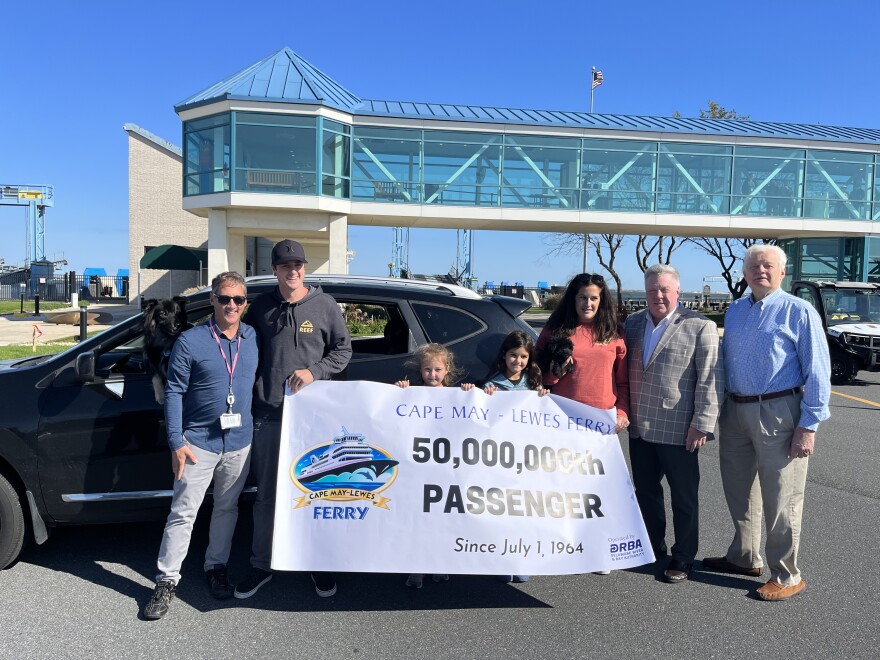 Five individuals along with two dogs hold a banner saying "Cape May-Lewes Ferry 50,000,000th Passenger" in front of a ferry port.