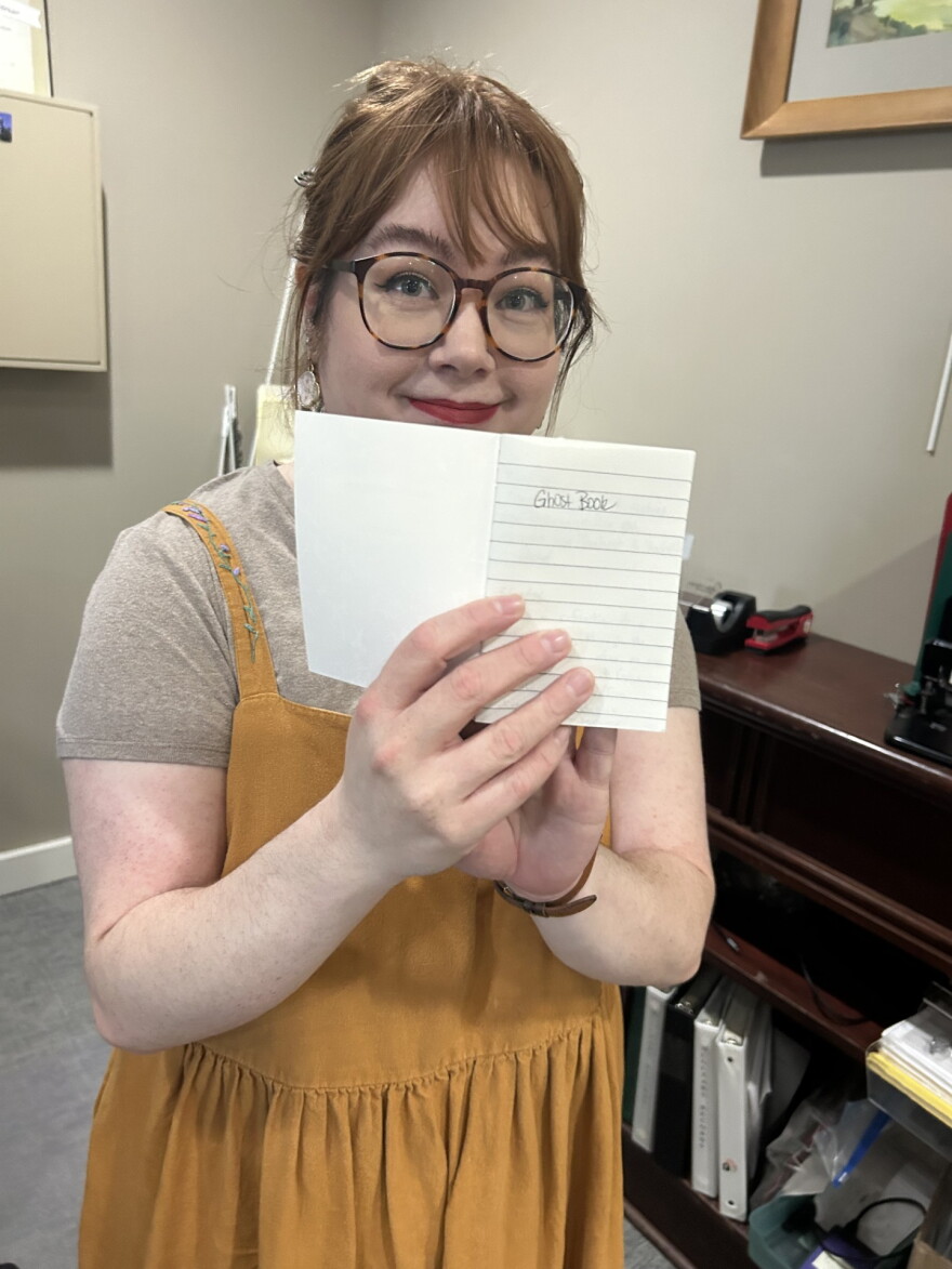 A woman in a yellow dress holds up a small notebook opened to a page that says, "Ghost Book."