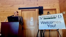 A man stands in front of a podium. A sign reading "Springfield City Club welcomes you" is to the right.