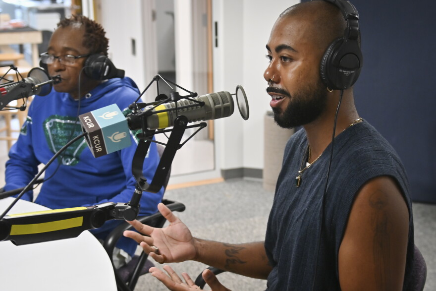 Two people sit inside a radio studio. The person at right is gesturing with both hands and talking at a microphone. The other appears to be listening.