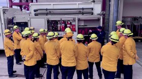 A group of people in yellow jackets and hard hats listen as a man points to components of a fire truck. 