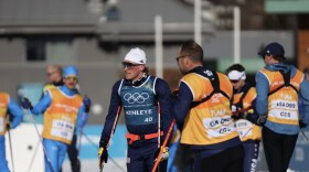 An Olympic cross-country skier wearing sunglasses and spandex stands on alongside team staff.
