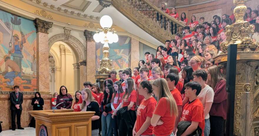 Teens in matching red shirts stand behind a woman who is speaking at a podium. 