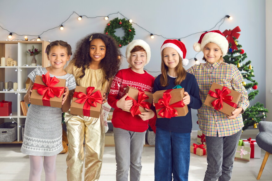 Group of happy smiling diverse children holding Christmas presents tied with beautiful red bows and looking at camera standing in cozy festively decorated living-room during fun party at home