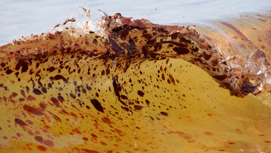 A light yellow ocean wave is dotted with blots of oil.