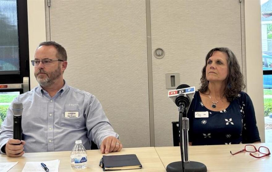 A man with a beard, wearing glasses, holds a microphone in his hand, and looks toward an audience, while he's seated behind a table. To his right, a woman looks in the same direction, while she's seated behind a microphone. A pair of red glasses lies on the table.