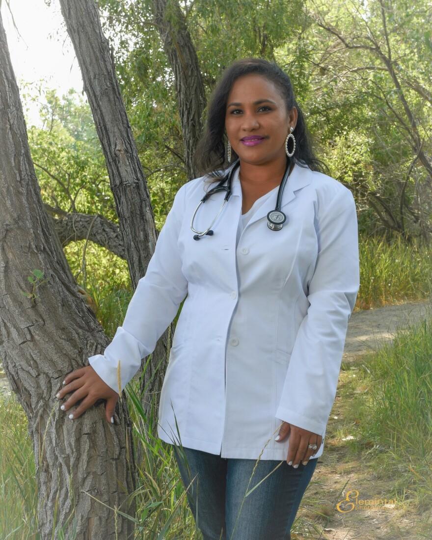 Salwa Mourtada Bamba, a Black woman with straightened, black hair, poses in front of a tree on a walking trail, greenery behind her. She is wearing a short doctor's coat and a stethoscope.