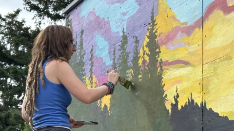 A woman wearing a blue tank top painting a tree on a giant mural.