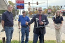 A man in a blazer speaks at a podium with two microphones, flanked by a man in a blue polo, an older man with a cane, and a woman in a black polo, with a gas station price sign in the background.