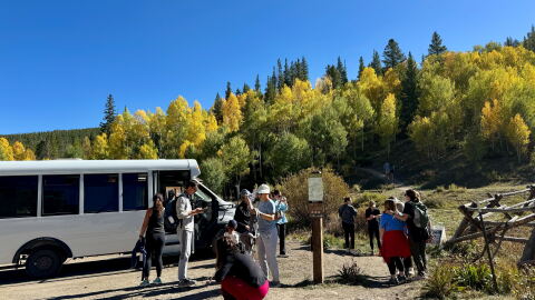 A shuttle bus drops off hikers in a dirt parking lot in front of a trail with yellow leaves