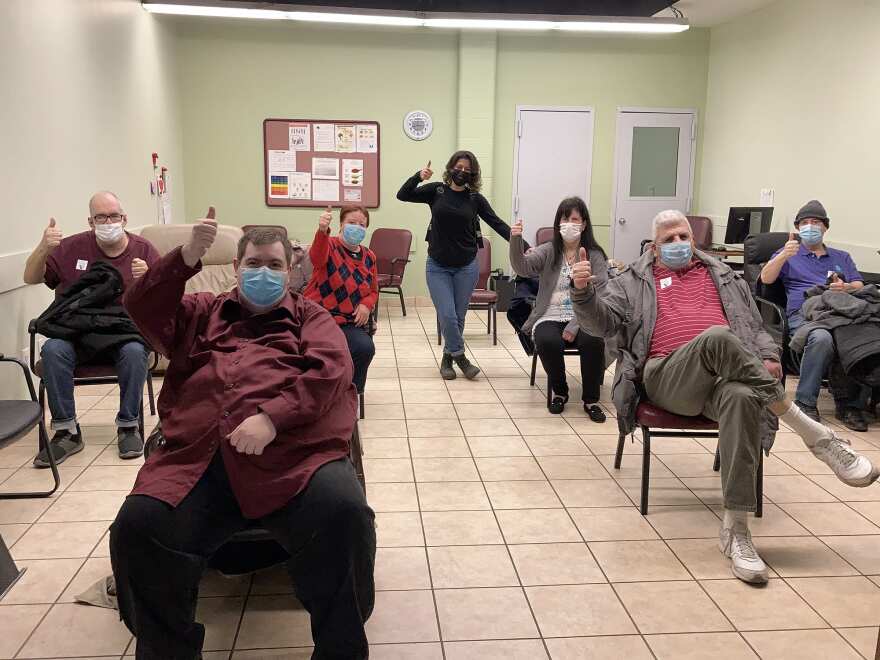 A group of six individuals sit in chairs with space between them, everyone giving a thumbs up. A staff member stands towards the back doing the same. Everyone is wearing a mask. They are in a green room with tile floors.