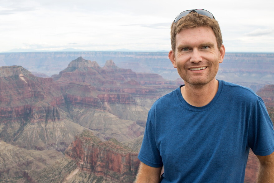 A man with brown hair wears a blue shirt and stands in front of a canyon. He smiles at the camera