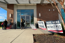 Photo of the outside of a  town hall. There are several signs saying "vote here" for different districts. One person is walking inside while two people are standing near a ballot drop box.
