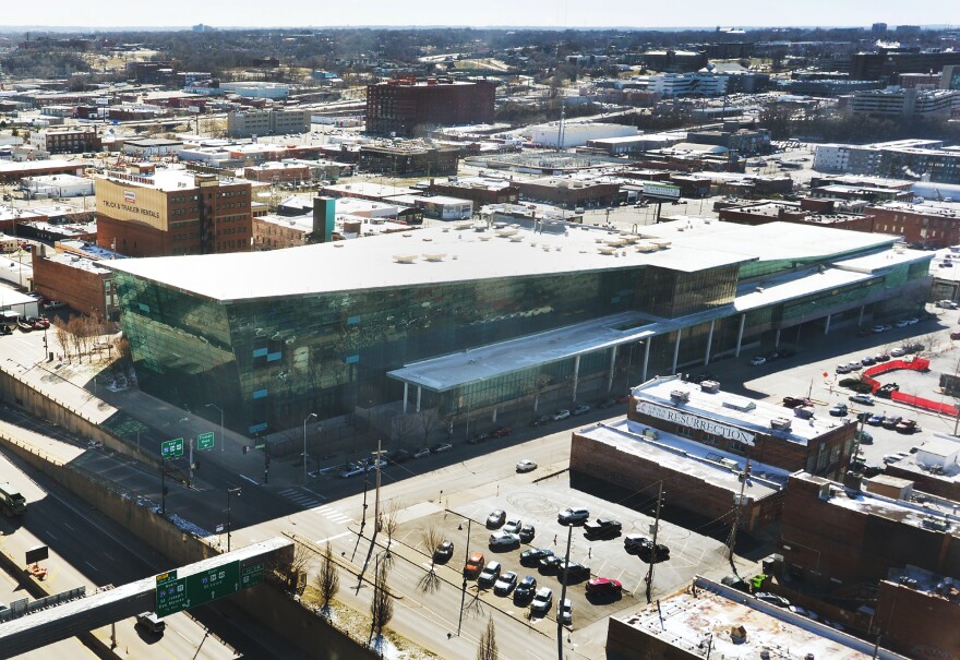 A large building with a green, glass exterior sits in the middle of a city block, surrounded by other smaller buildings.